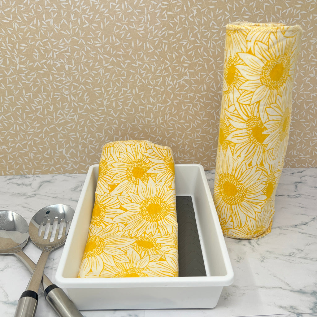 Flannel reusable paper towels. This pictures shows possible display options on Kitchen counter. One group is folded in a basket and the other group rolled on a cylinder. Cotton fabric has a large overlapping  sketch of yellow daisies 