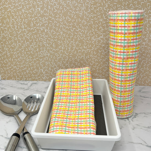 Flannel reusable paper towels. This pictures shows possible display options on Kitchen counter. One group is folded in a basket and the other group rolled on a cylinder. place setting. Cotton fabric has a yellow, turquoise and light red plaid pattern on a white background