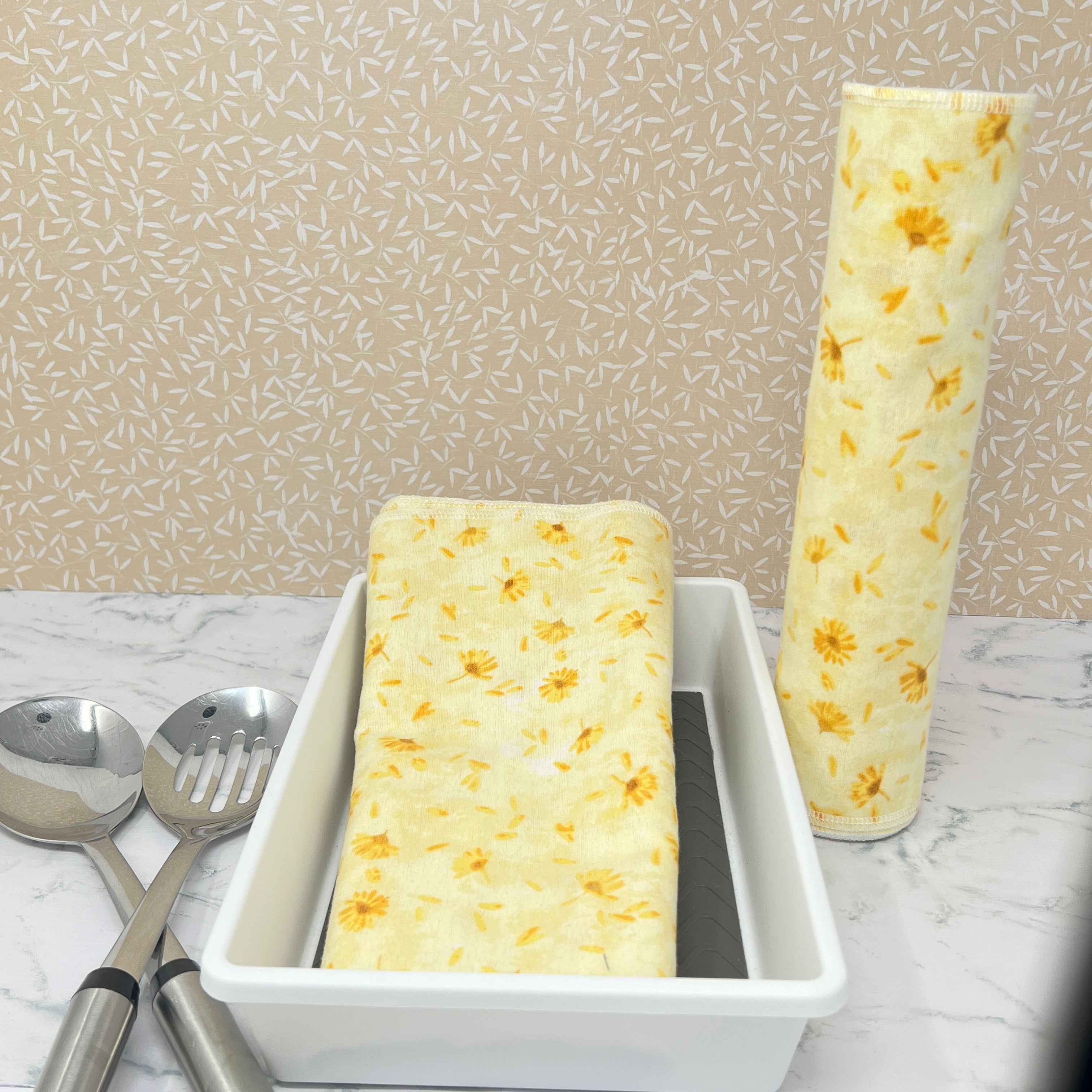 Flannel reusable paper towels. This pictures shows possible display options on Kitchen counter. One group is folded in a basket and the other group rolled on a cylinder. place setting. Cotton fabric has yellow and gold dandelions  on a soft yellow on background