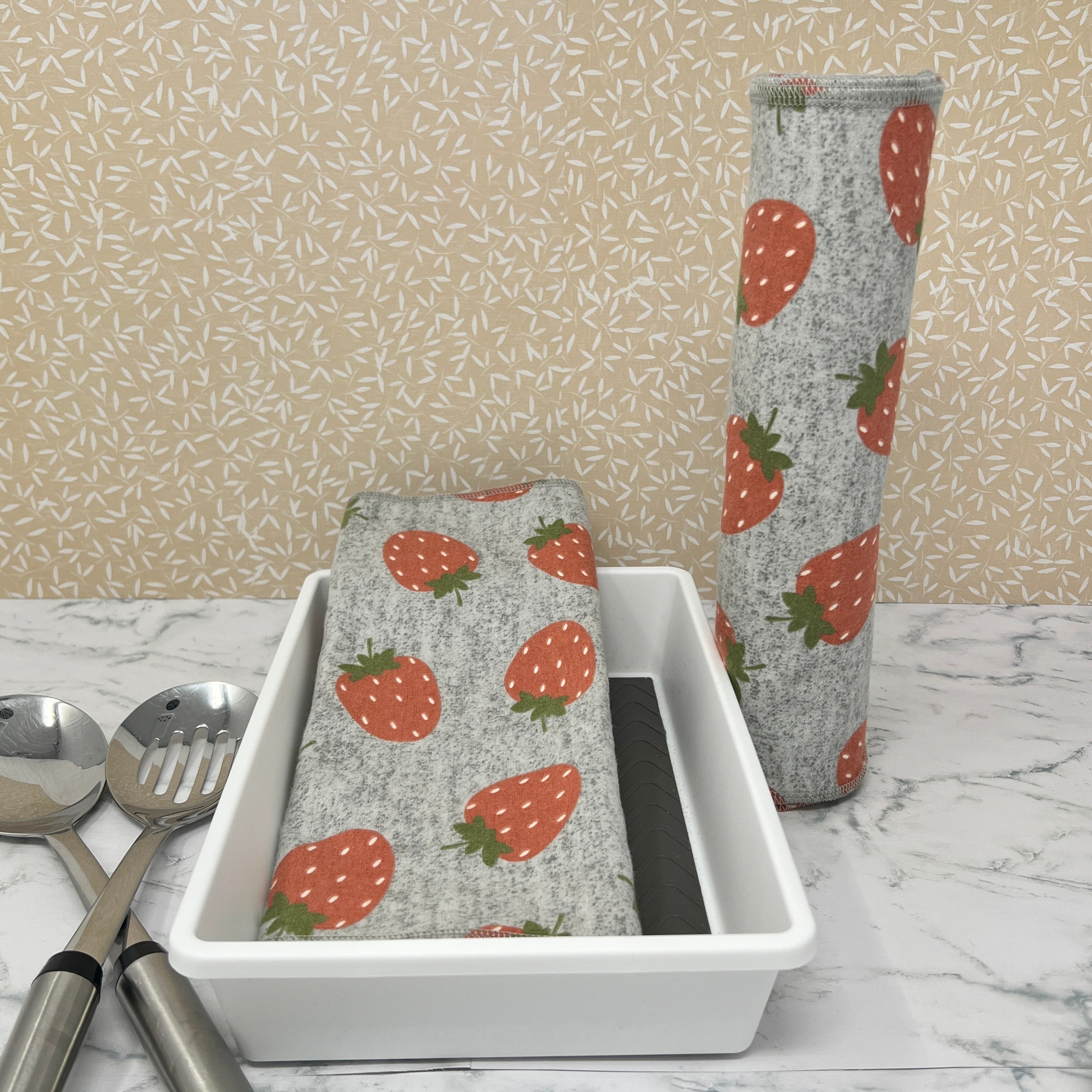 Flannel reusable paper towels. This pictures shows possible display options on Kitchen counter. One group is folded in a basket and the other group rolled on a cylinder. Cotton fabric has random red strawberries on a heather gray background