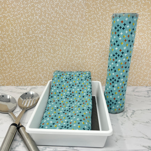 Flannel reusable paper towels  showing possible counter displays with a group folded in a basket and another group rolled onto a cylinder as possible . Fabric is a turquoise blue background with stars or crosshatches in blue, yellow, red , white and black, in a scattered repeating pattern. 