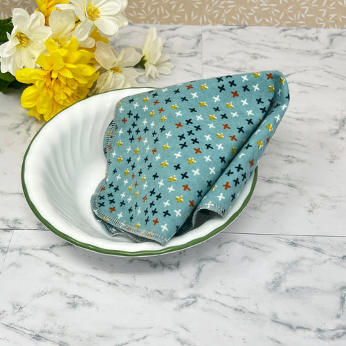 Flannel reusable paper towels. this pictures shows on towel in a place setting. Fabric is a turquoise blue background with stars or crosshatches in blue, yellow, red , white and black, in a scattered repeating pattern. 