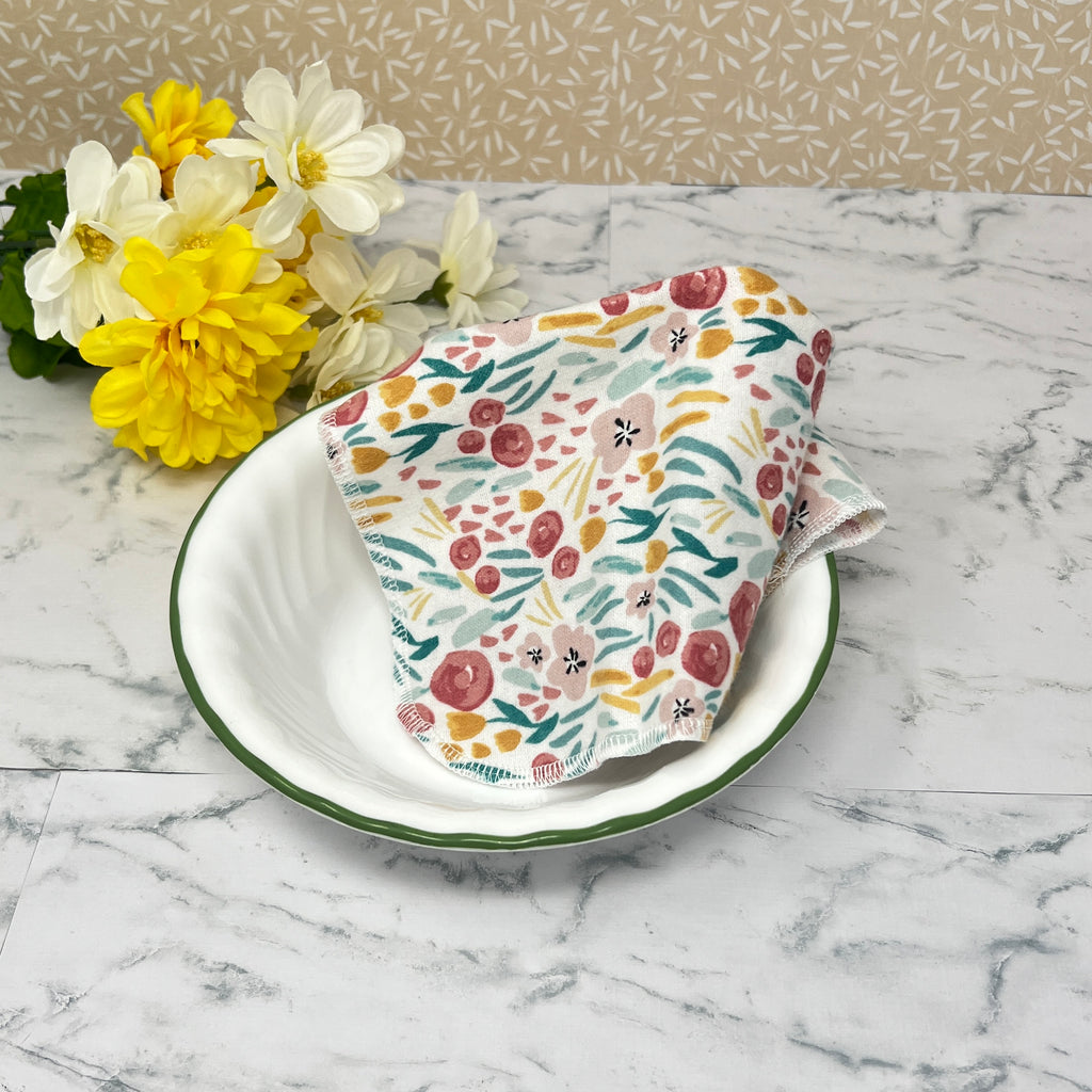 Flannel reusable paper towels. this pictures shows one towel in a place setting.   Fabric has a floral and greenery  pattern in a painted "style" with red and gold flowers, and greenery on a white background.