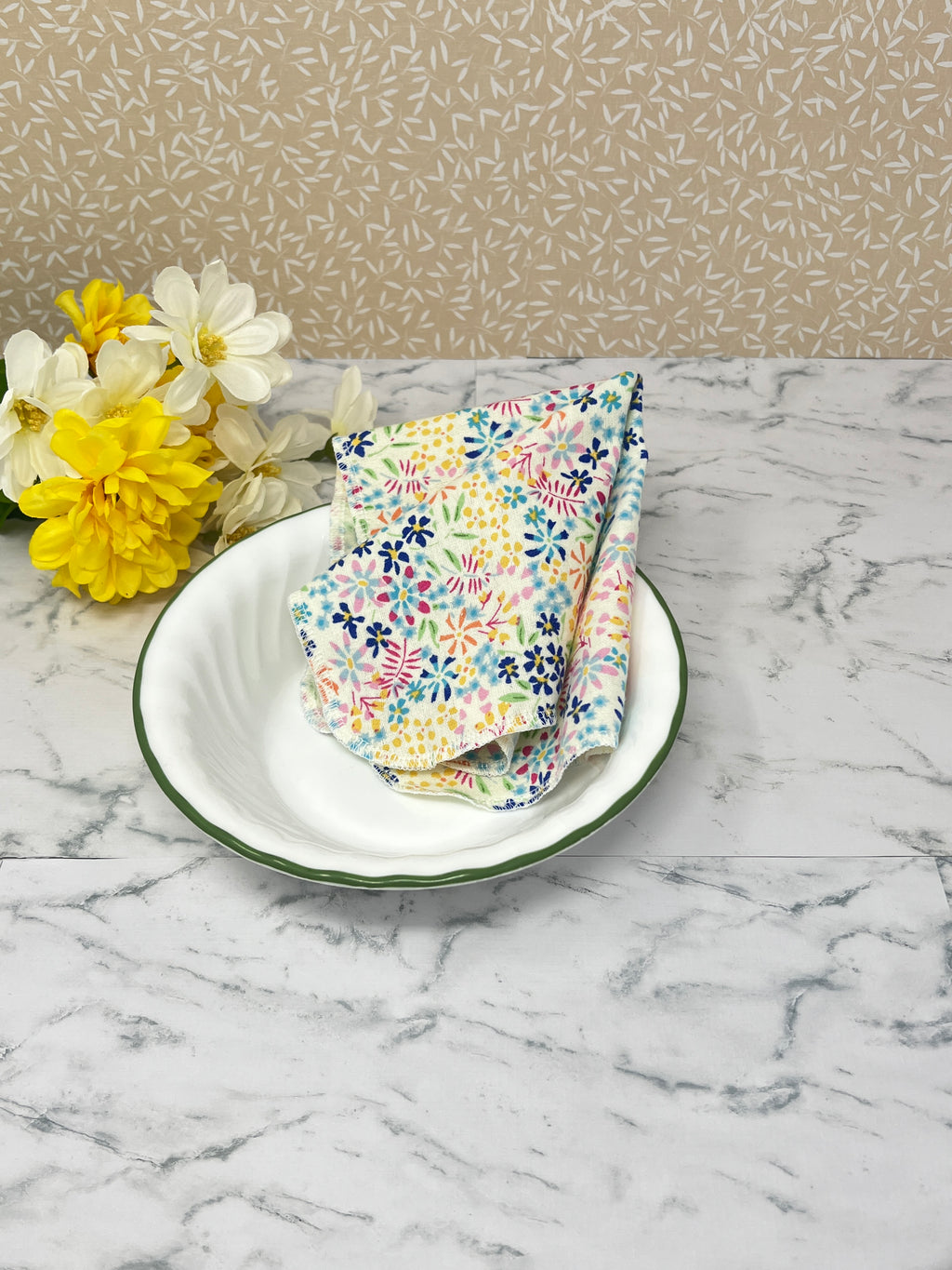 Flannel reusable paper towels. this pictures shows one towel in a place setting.   Cotton fabric has a ditzy floral print pattern of tiny colorful flowers on a white background.