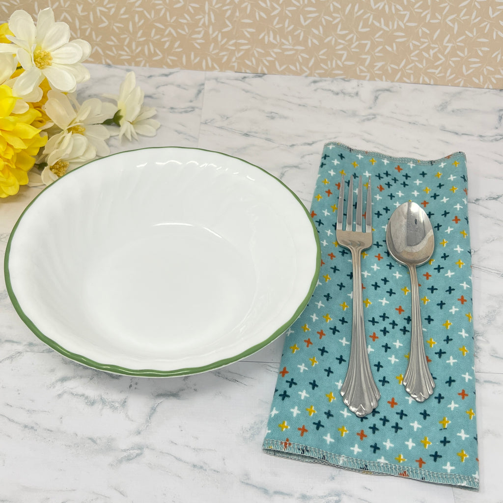 Flannel reusable paper towels. this pictures shows one towel in a place setting. Fabric is a turquoise blue background with stars or crosshatches in blue, yellow, red , white and black, in a scattered repeating pattern. 