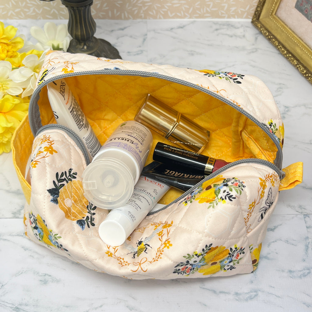 A large quilted  boxy bag with a zipper top and handle. Also known as a cosmetic or makeup bag, and toiletry pouch.  Fabric is cream color background with yellow and black honey bees, bee hive, and florals in a repeating pattern.  Picture displays the yellow interior lining and  shows an assortment of cosmetics as a visual possible use of bag. 