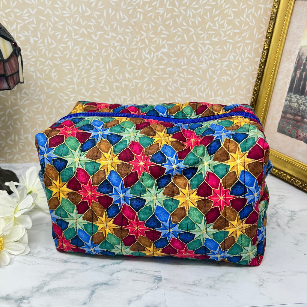 A side view of a large quilted makeup bag or cosmetic bag pouch also known as a toiletry bag,  with zipper and handle. Fabric is cotton and has a colorful array of kaleidoscope shapes in varying shades of  blue, red, green and yellow. A coordinating blue handle to match the blue interior lining