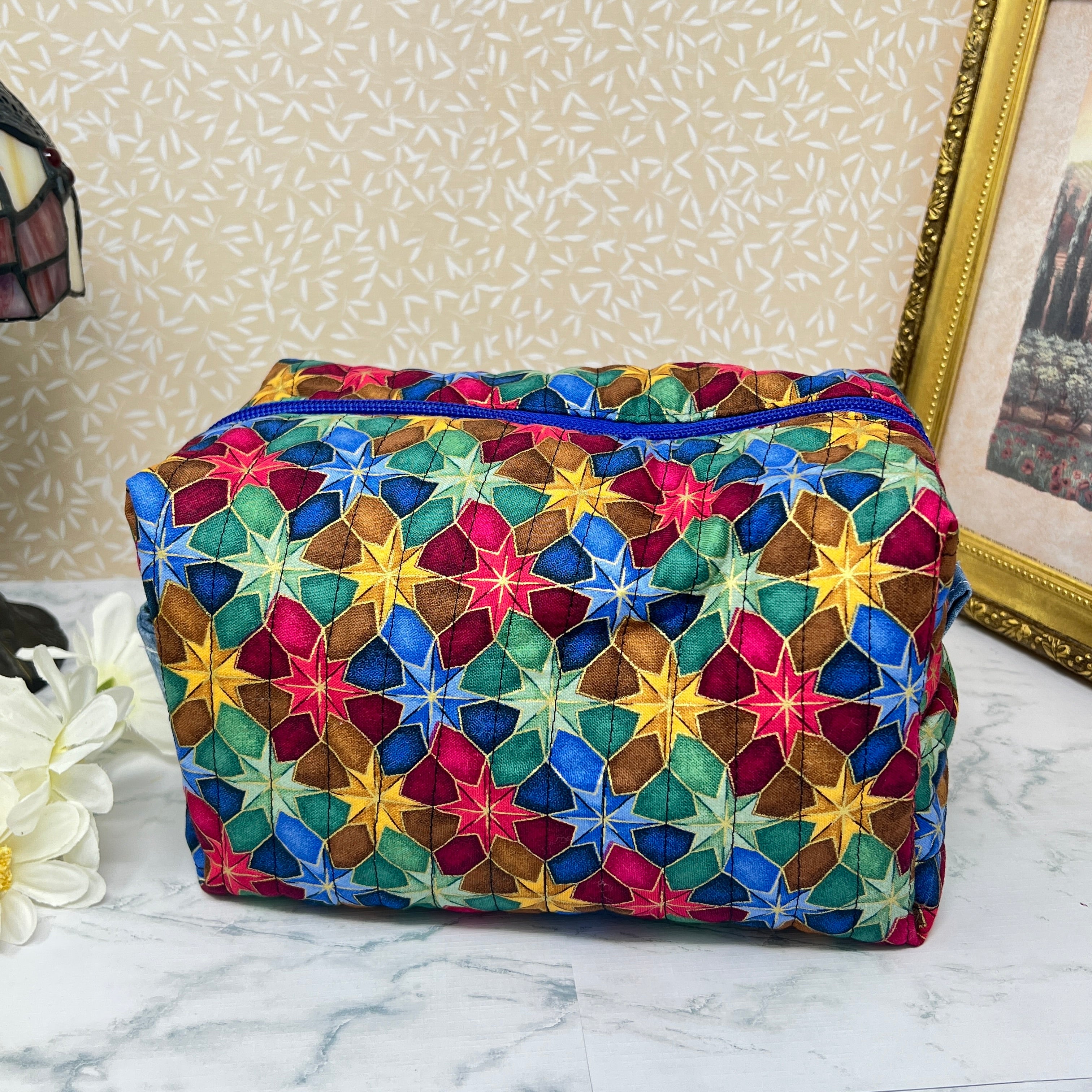 A side view of a large quilted makeup bag or cosmetic bag pouch also known as a toiletry bag,  with zipper and handle. Fabric is cotton and has a colorful array of kaleidoscope shapes in varying shades of  blue, red, green and yellow. A coordinating blue handle to match the blue interior lining
