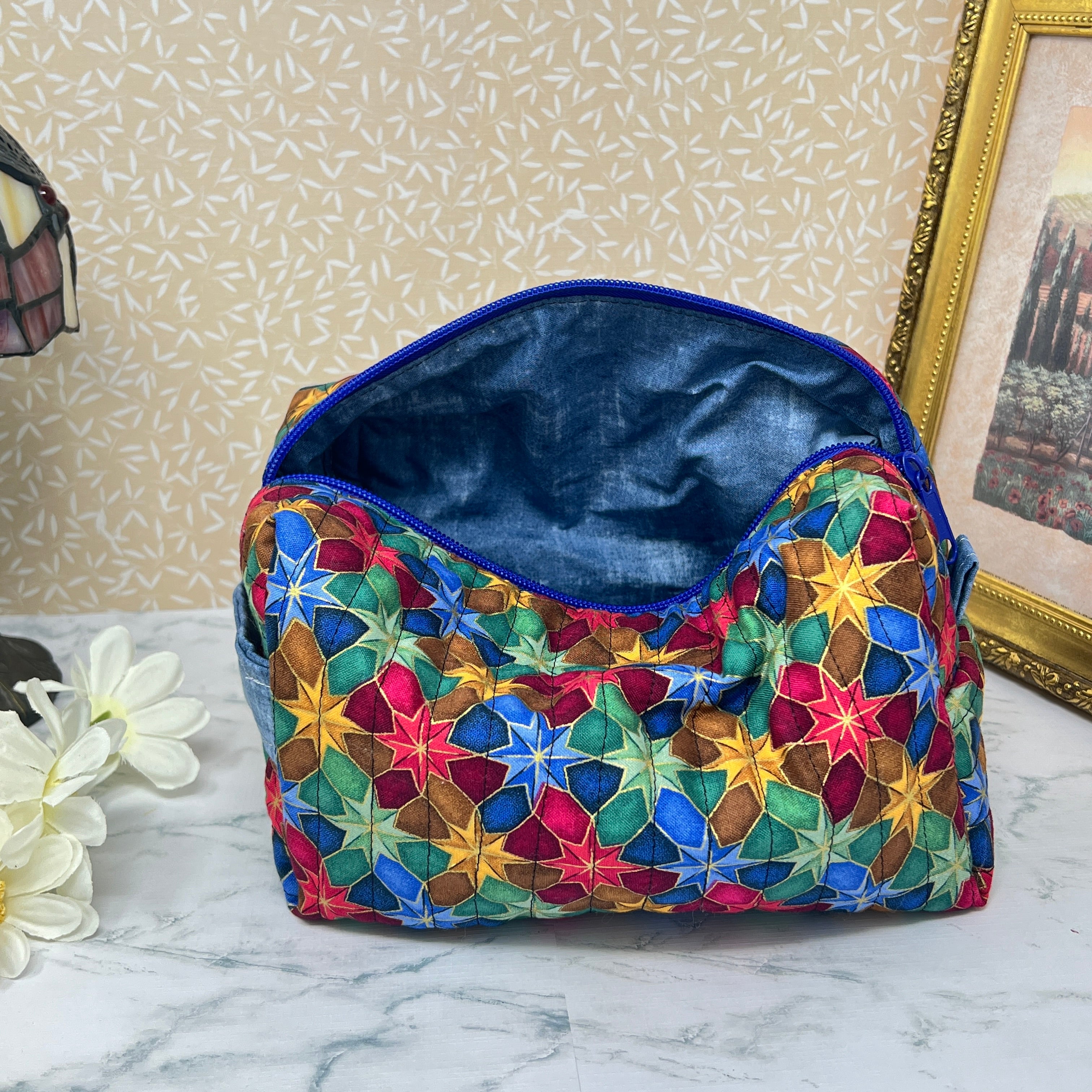 A large quilted makeup bag or cosmetic bag pouch also known as a toiletry bag,  with zipper and handle. Fabric is cotton and has a colorful array of kaleidoscope shapes in varying shades of  blue, red, green and yellow. A coordinating blue handle to match the blue interior lining