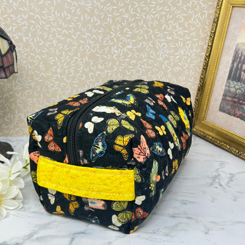 A large quilted cosmetic bag with zipper and handle. Also known as a makeup bag or toiletry bag. Fabric is a colorful assortment of butterflies and feathers on a black background with a yellow handle.