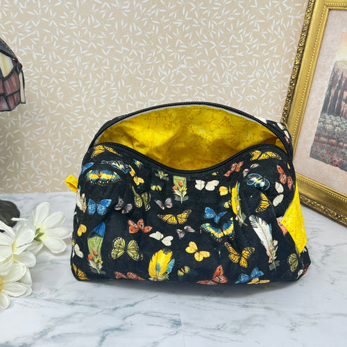 A large quilted cosmetic bag with zipper and handle showing the yellow interior lining . Also known as a makeup bag or toiletry bag or pouch. Fabric is a colorful assortment of butterflies and feathers on a black background with a yellow handle.