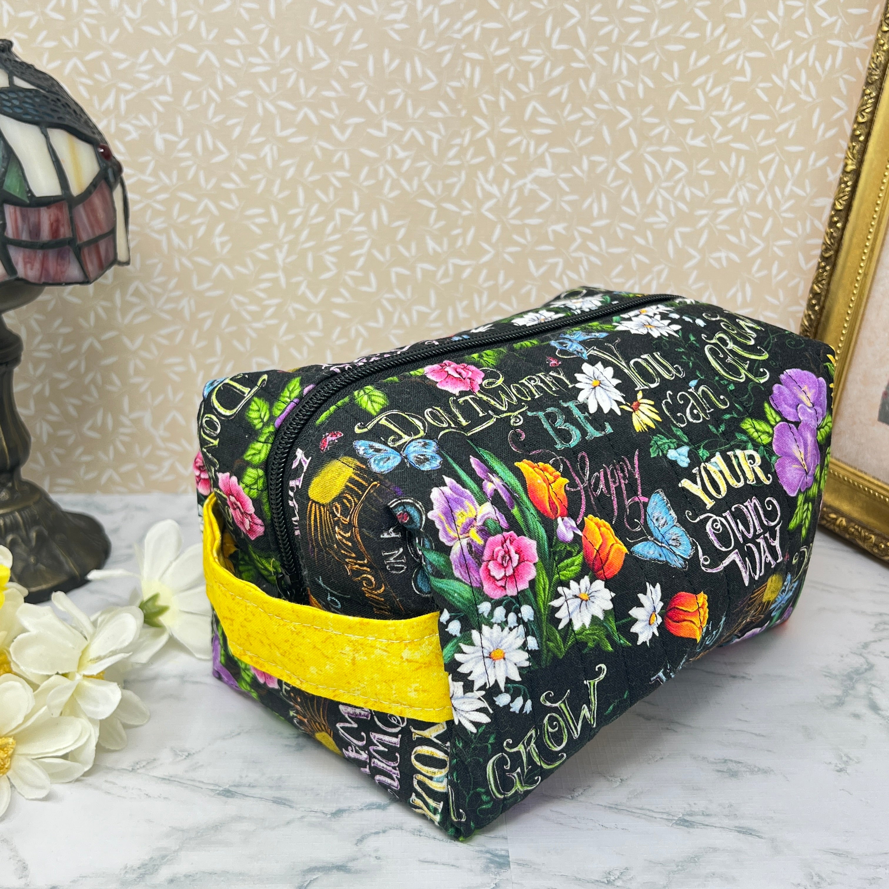 A large quilted boxy bag with zipper and handle. Fabric is colorful flowers and positive affirmation text on a black background. Yellow handle and yellow liner