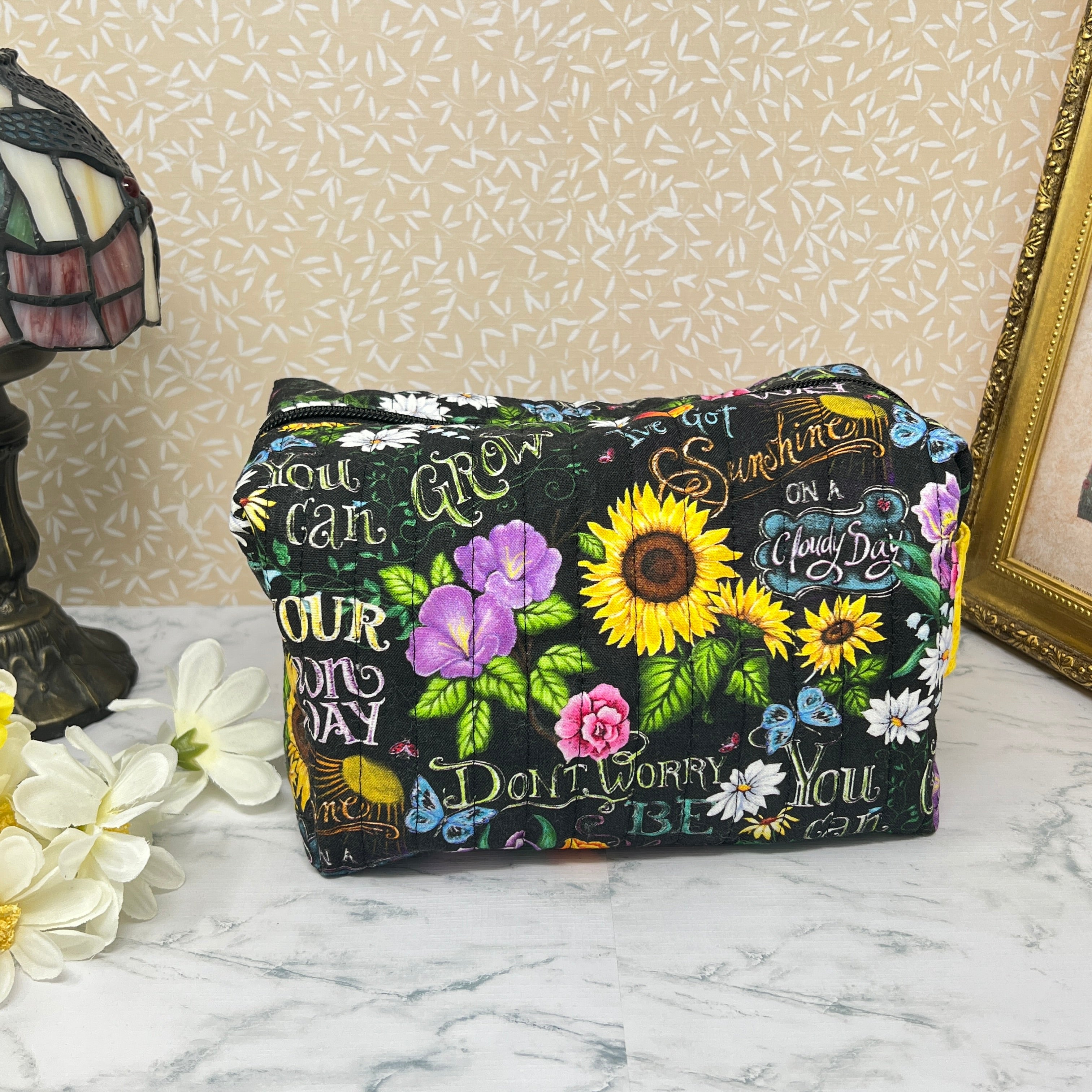 A large quilted boxy bag with zipper and handle. Fabric is colorful flowers and positive affirmation text on a black background. Yellow handle and yellow liner. Text reads; Don't worry be happy, Sunshine on a cloudy day, You can grow your own way