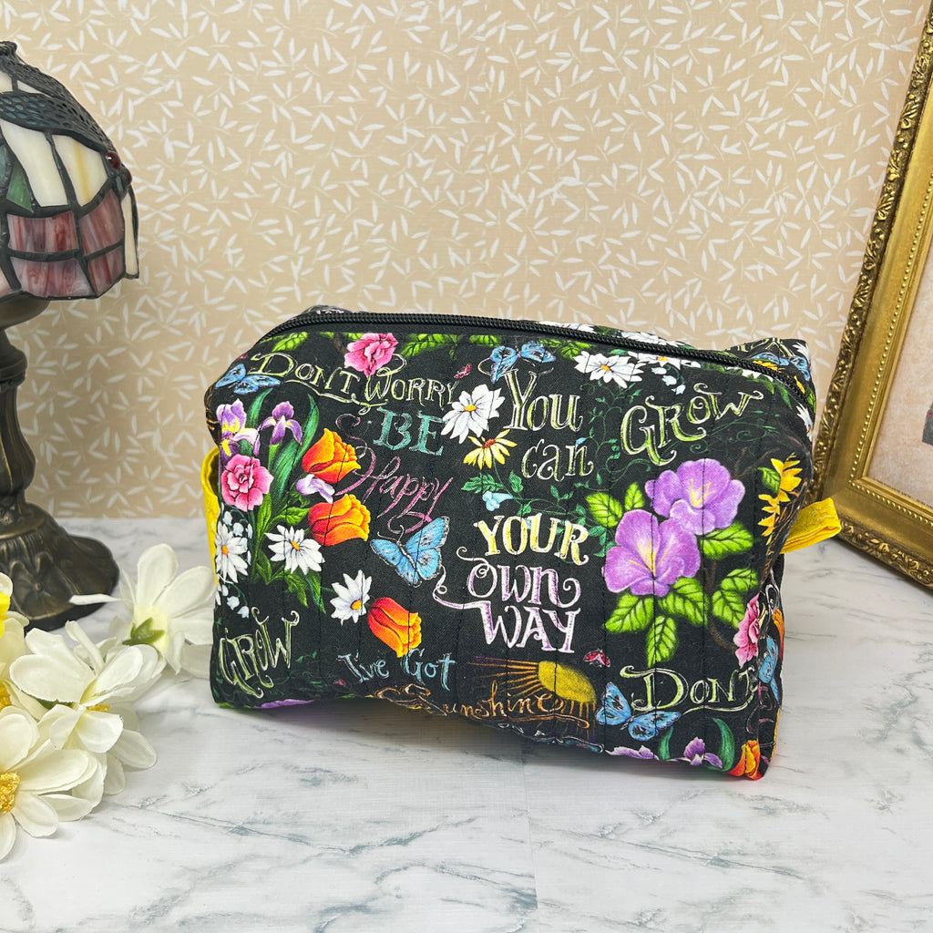 A large quilted boxy bag with zipper and handle. Fabric is colorful flowers and positive affirmation text on a black background. Yellow handle and yellow liner. Text reads; Don't worry be happy, Sunshine on a cloudy day, You can grow your own way