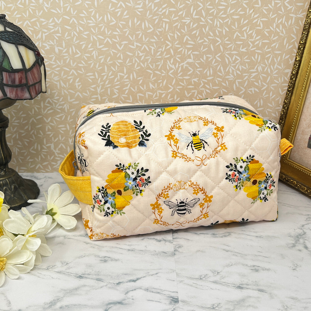 A large quilted pouch with a zipper top and handle. Fabric is cream color with yellow and black honey bees and a bee hive. Side view