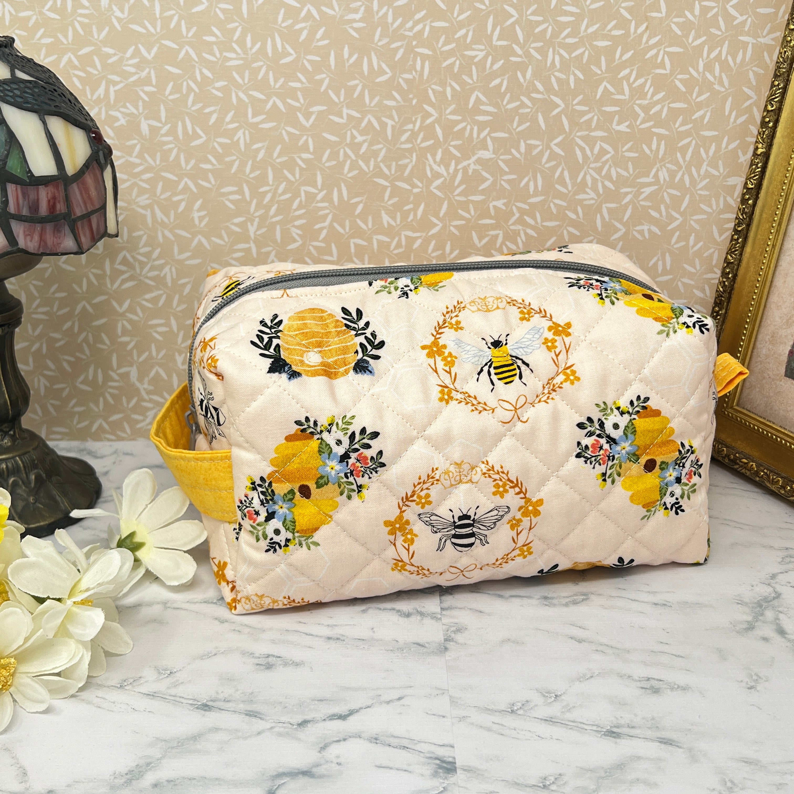 A large quilted pouch with a zipper top and handle. Fabric is cream color with yellow and black honey bees and a bee hive. Side view