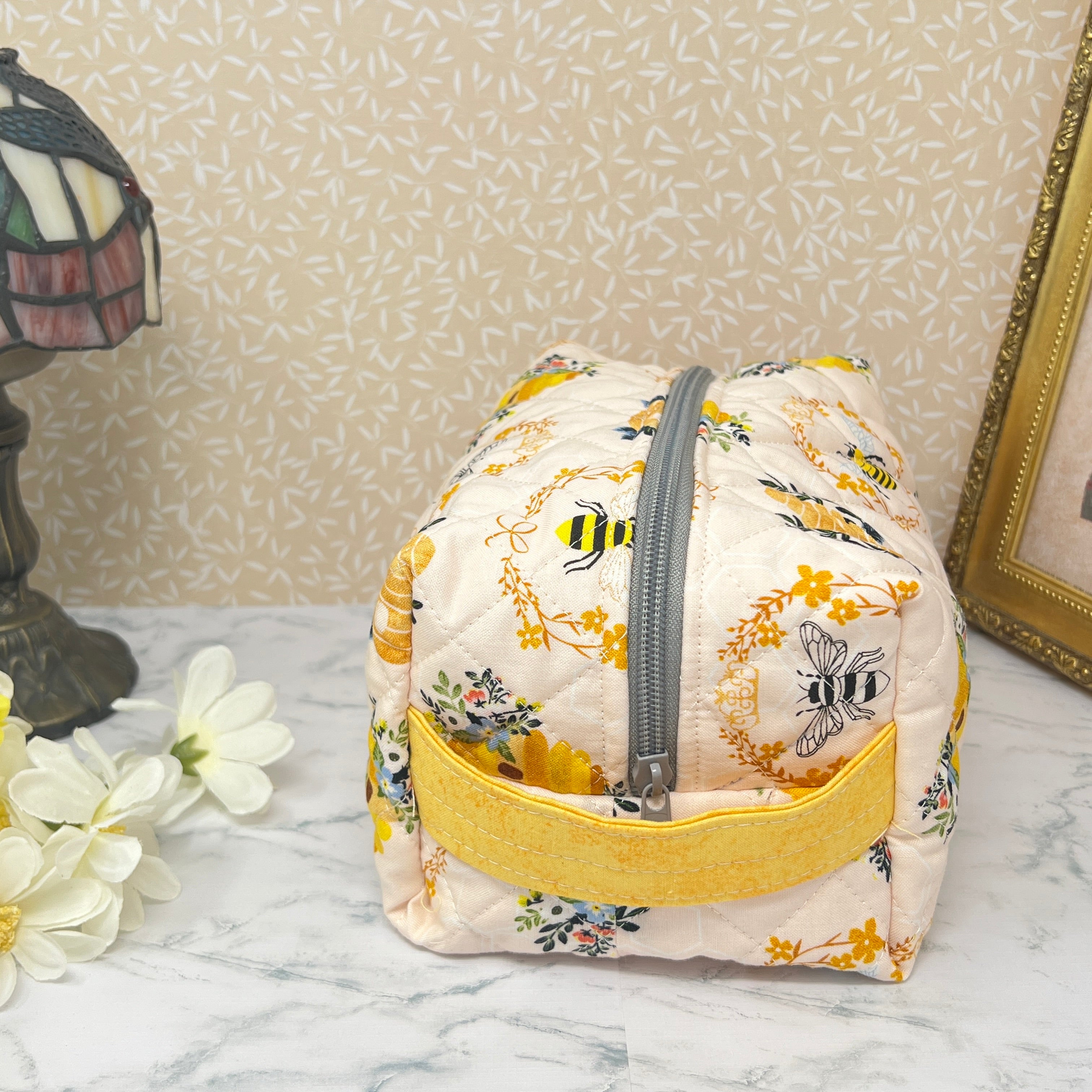 A large quilted pouch with a zipper top and handle. Fabric is cream color with yellow and black honey bees and a bee hive. End view showing yellow handle