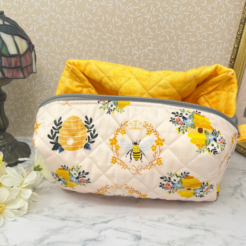 A large quilted  boxy bag with a zipper top and handle. Also known as a cosmetic or makeup bag, and toiletry pouch.  Fabric is cream color background with yellow and black honey bees, bee hive, and florals in a repeating pattern. Handle and quilted interior lining are  yellow.