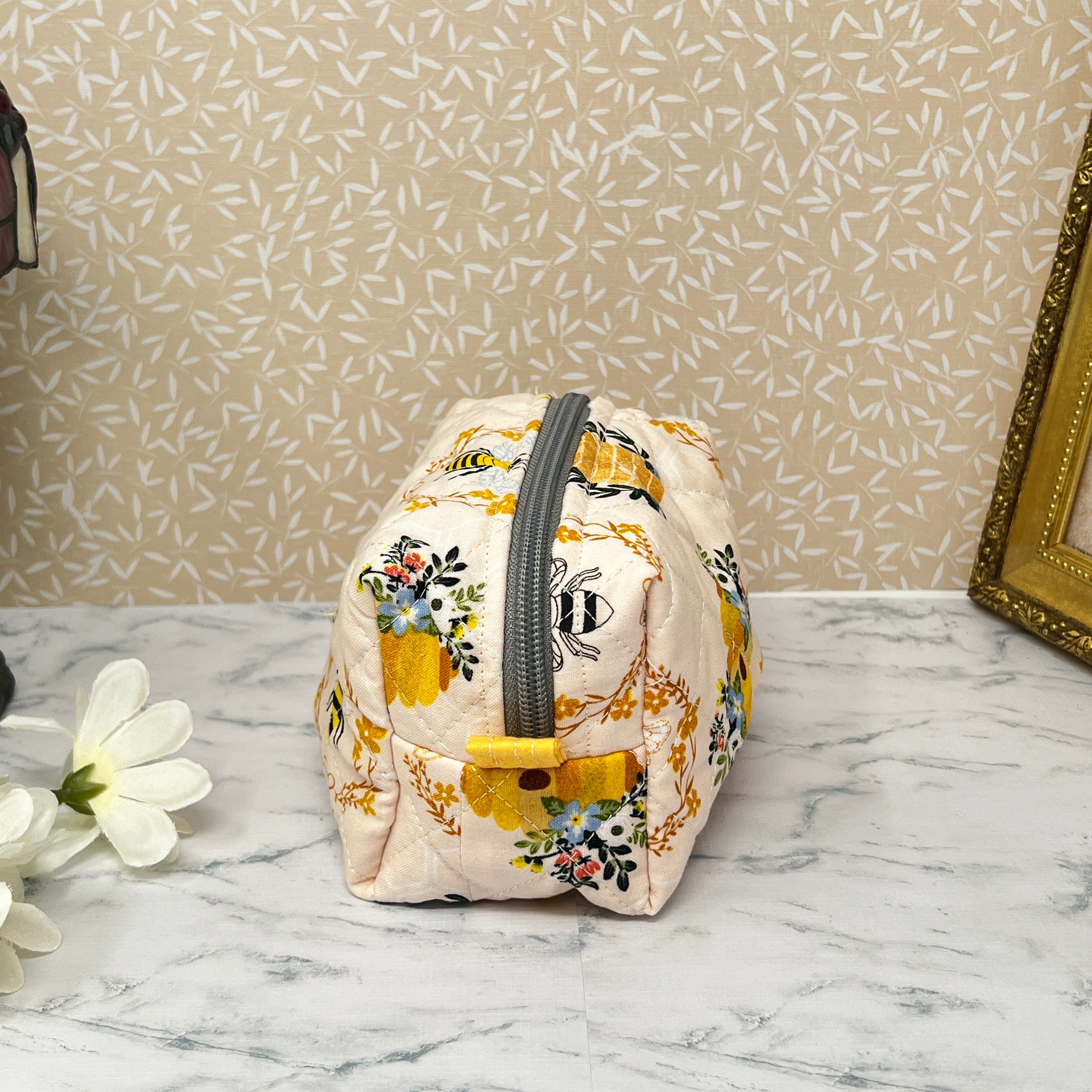 Small quilted pouch with a zipper top and handle. Fabric is cream color with honey bees and a bee hive. handle and lining are yellow