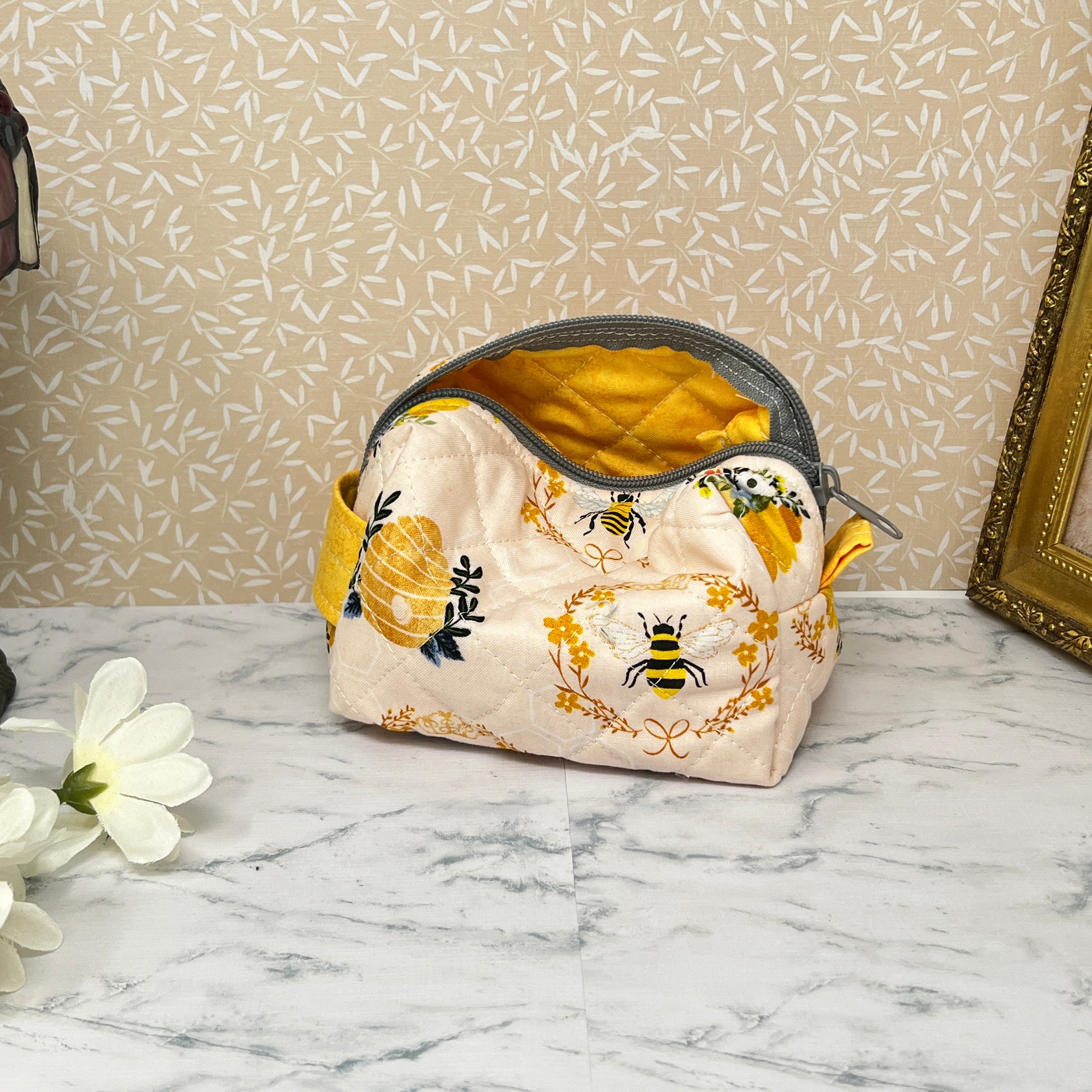 Small quilted pouch with a zipper top and handle. Fabric is cream color with honey bees and a bee hive. handle and lining are yellow. Showing yellow quilted interior of bag and yellow tab
