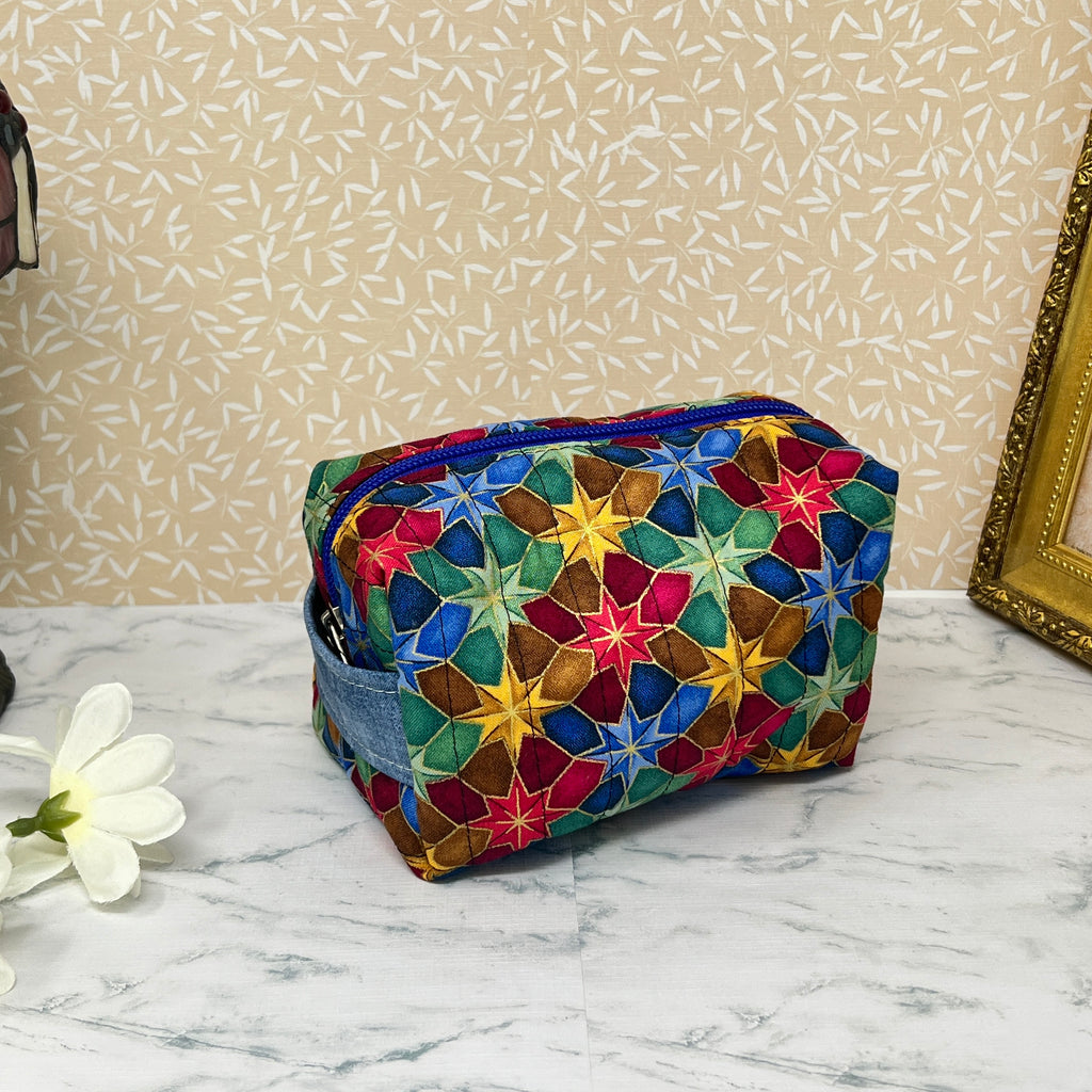 Small quilted makeup bag or cosmetic bag pouch with zipper and handle. Fabric is cotton and has a colorful array of kaleidoscope shapes in varying shades of  blue, red, green and yellow. Blue handle to match lining