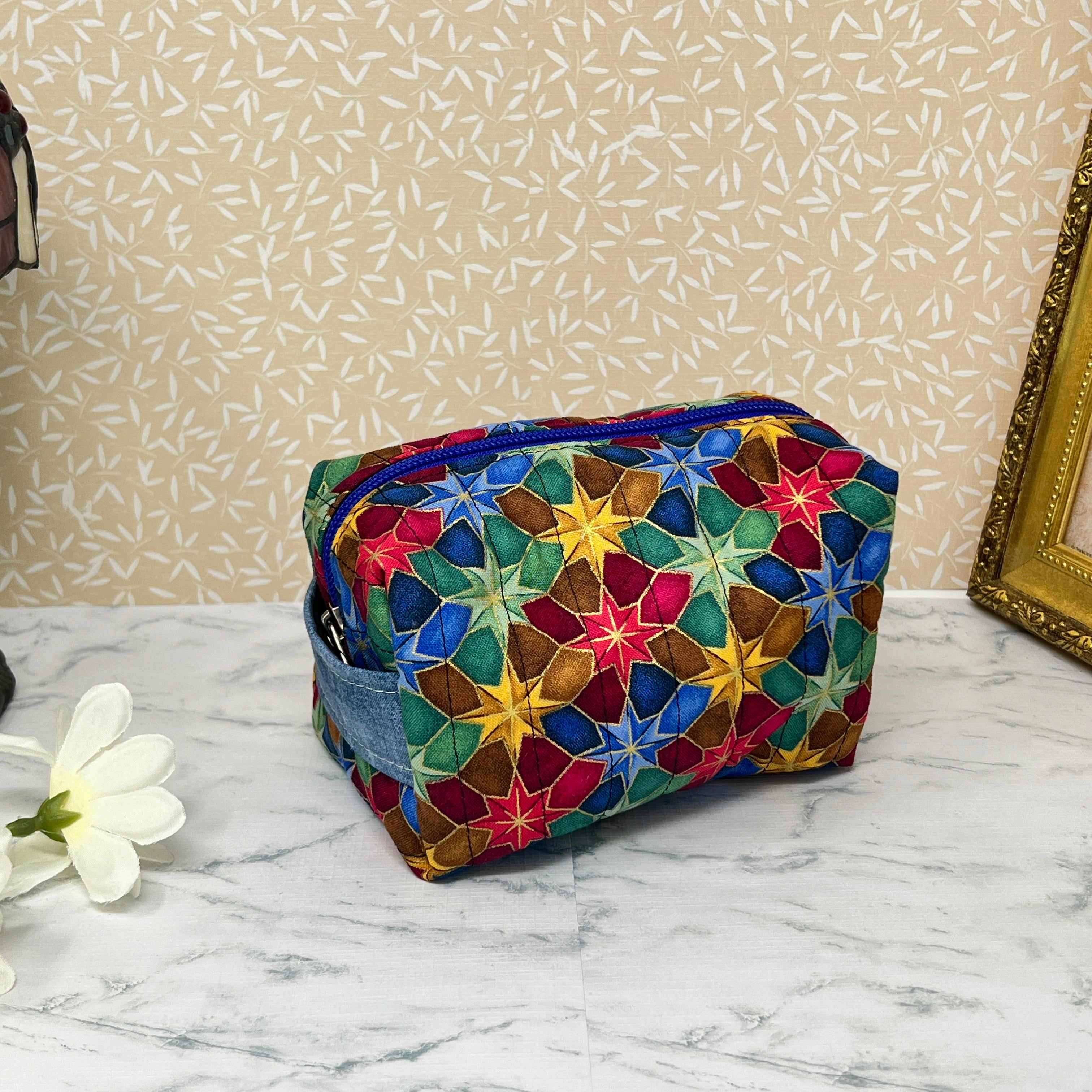 Small quilted makeup bag or cosmetic bag pouch with zipper and handle. Fabric is cotton and has a colorful array of kaleidoscope shapes in varying shades of  blue, red, green and yellow. Blue handle to match lining