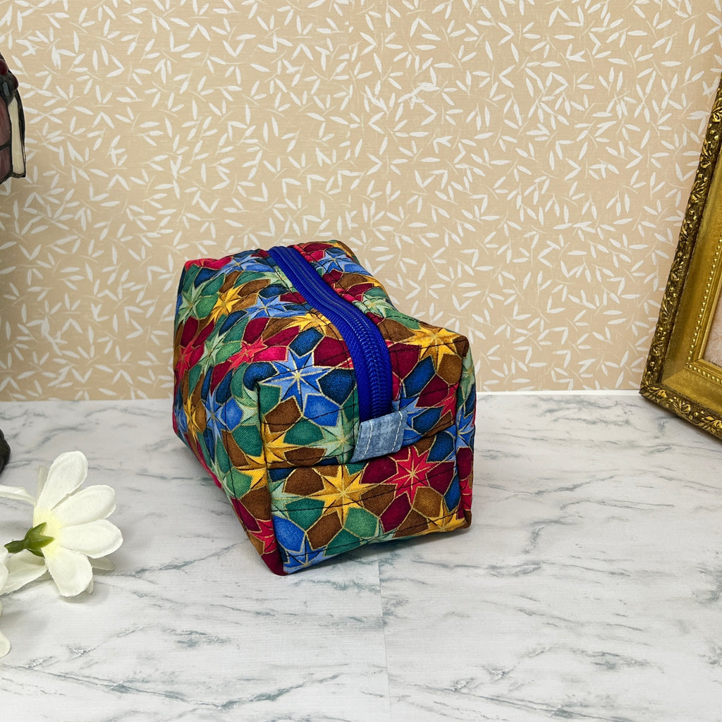 Small quilted makeup bag or cosmetic bag pouch also known as a toiletry bag,  with zipper and handle. Fabric is cotton and has a colorful array of kaleidoscope shapes in varying shades of  blue, red, green and yellow. Blue handle to match the blue lining