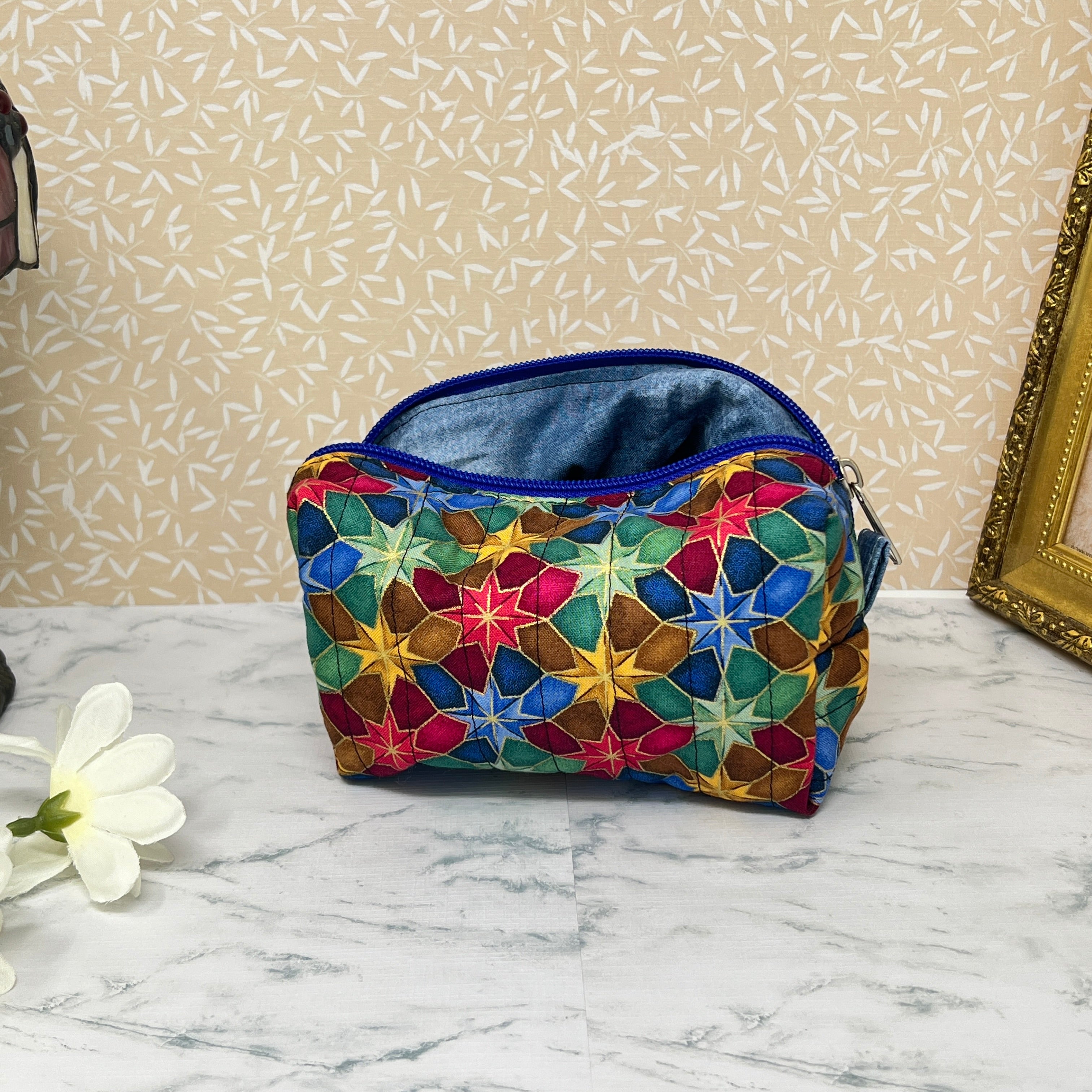 Small quilted makeup bag or cosmetic bag pouch with zipper and handle. Fabric is cotton and has a colorful array of kaleidoscope shapes in varying shades of  blue, red, green and yellow. Blue handle to match the blue lining