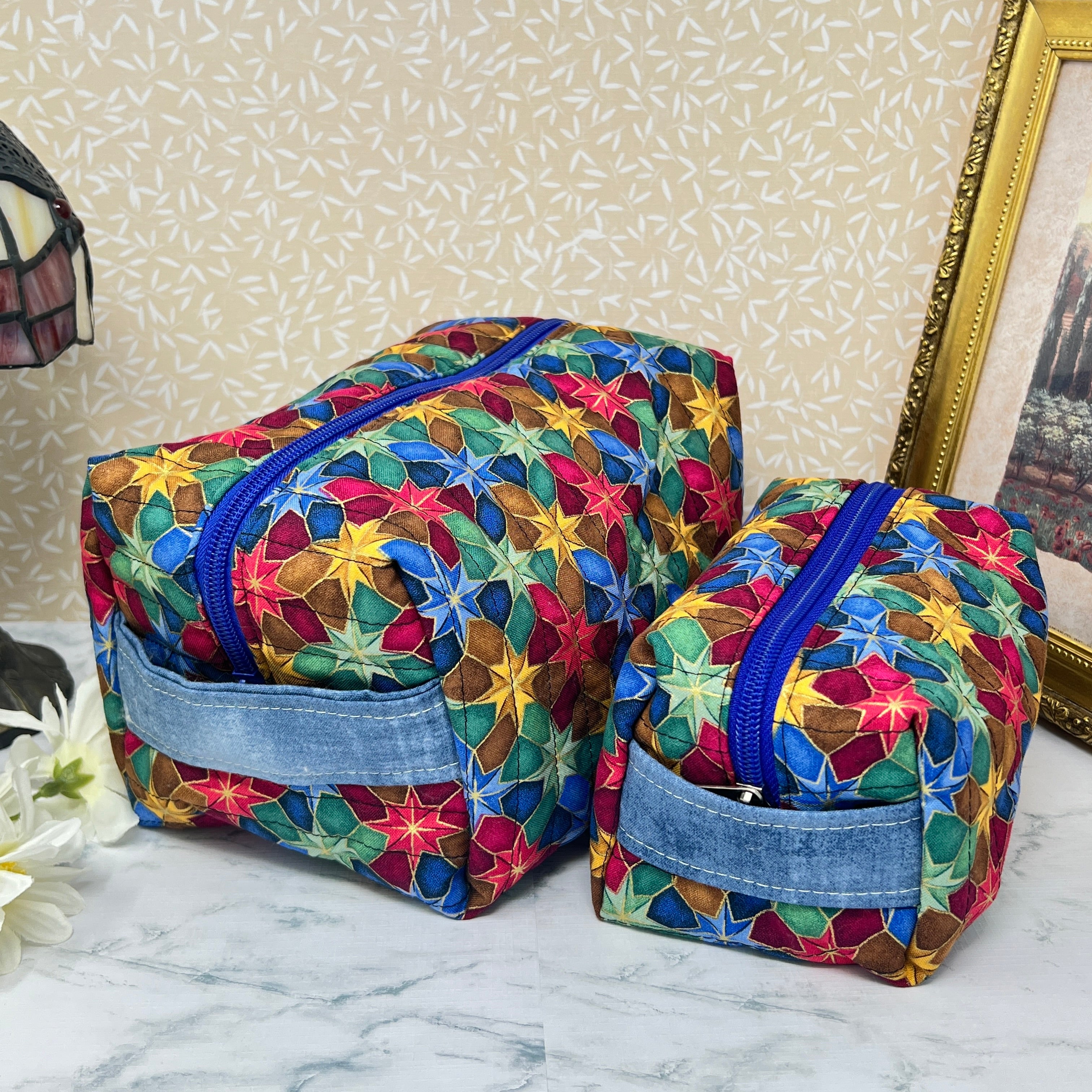Picture shows a large and a small quilted makeup bag or cosmetic bag pouch with zipper and handle. Fabric is cotton and has a colorful array of kaleidoscope shapes in varying shades of  blue, red, green and yellow. Blue handle to match the blue lining