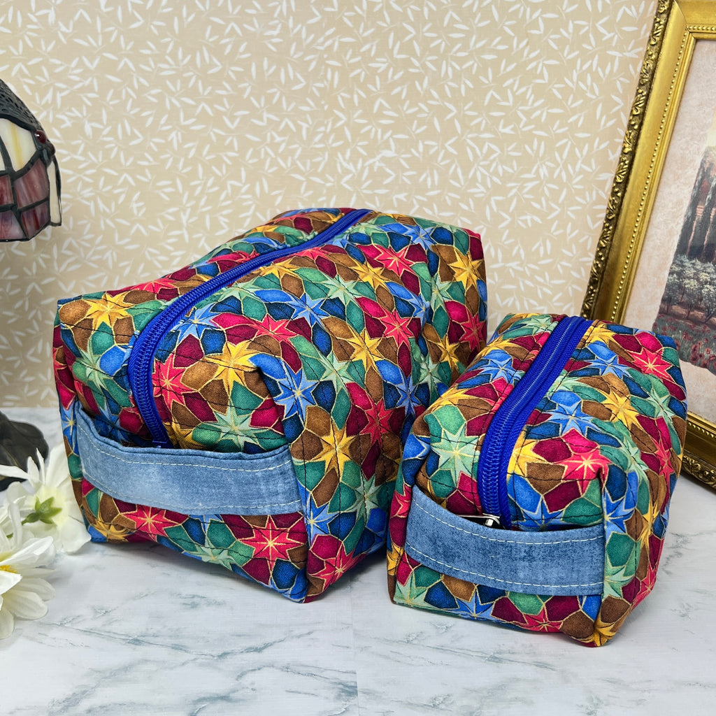 A large and a small quilted makeup bag or cosmetic bag pouch set, also known as a toiletry bags,  with zipper and handle. Fabric is cotton and has a colorful array of kaleidoscope shapes in varying shades of  blue, red, green and yellow. A coordinating blue handle to match the blue interior lining