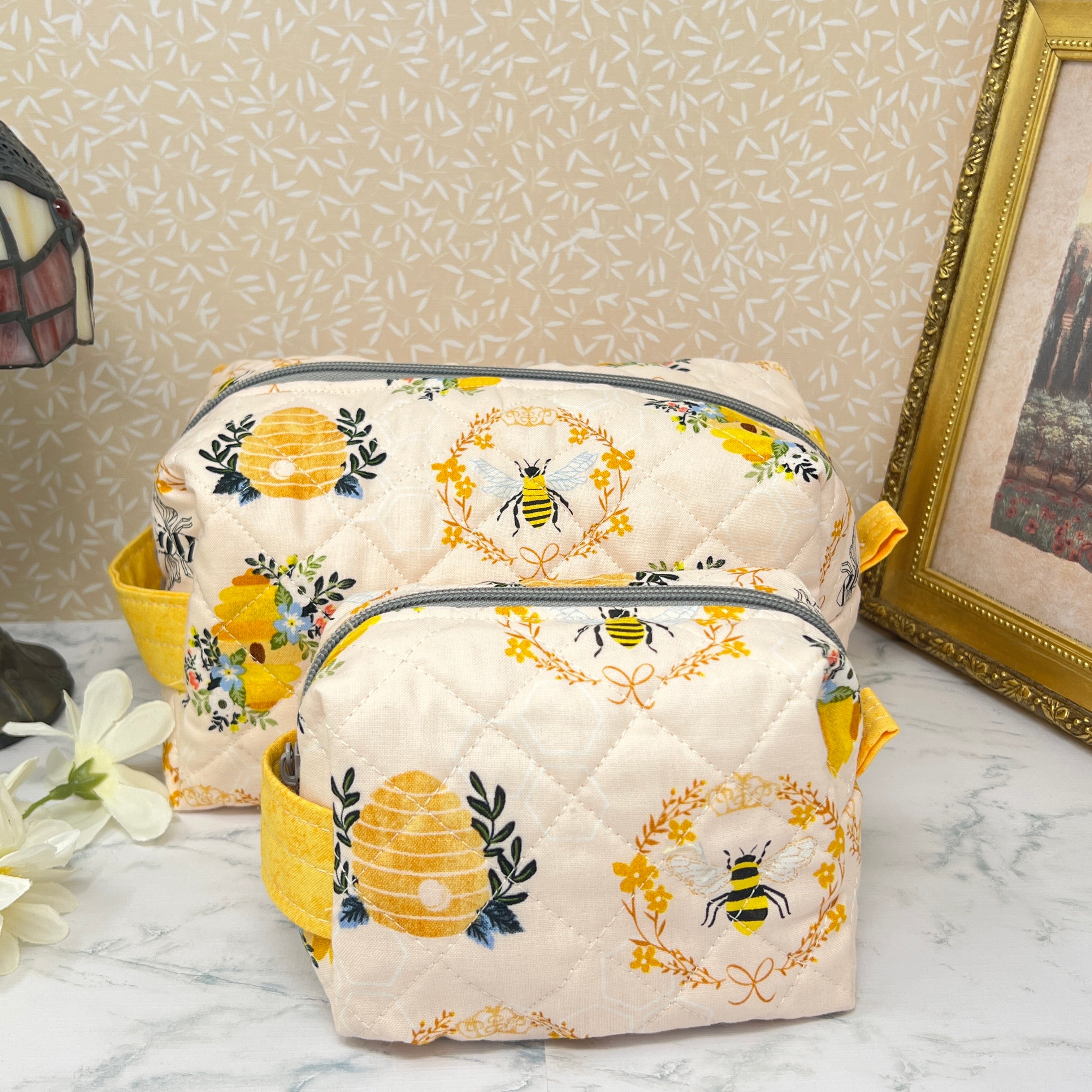 A large  and a small quilted  boxy bag  set with a zipper top and handle. Also known as a cosmetic or makeup bag, and toiletry pouch.  Fabric is cream color background with yellow and black honey bees, bee hive, and florals in a repeating pattern.  