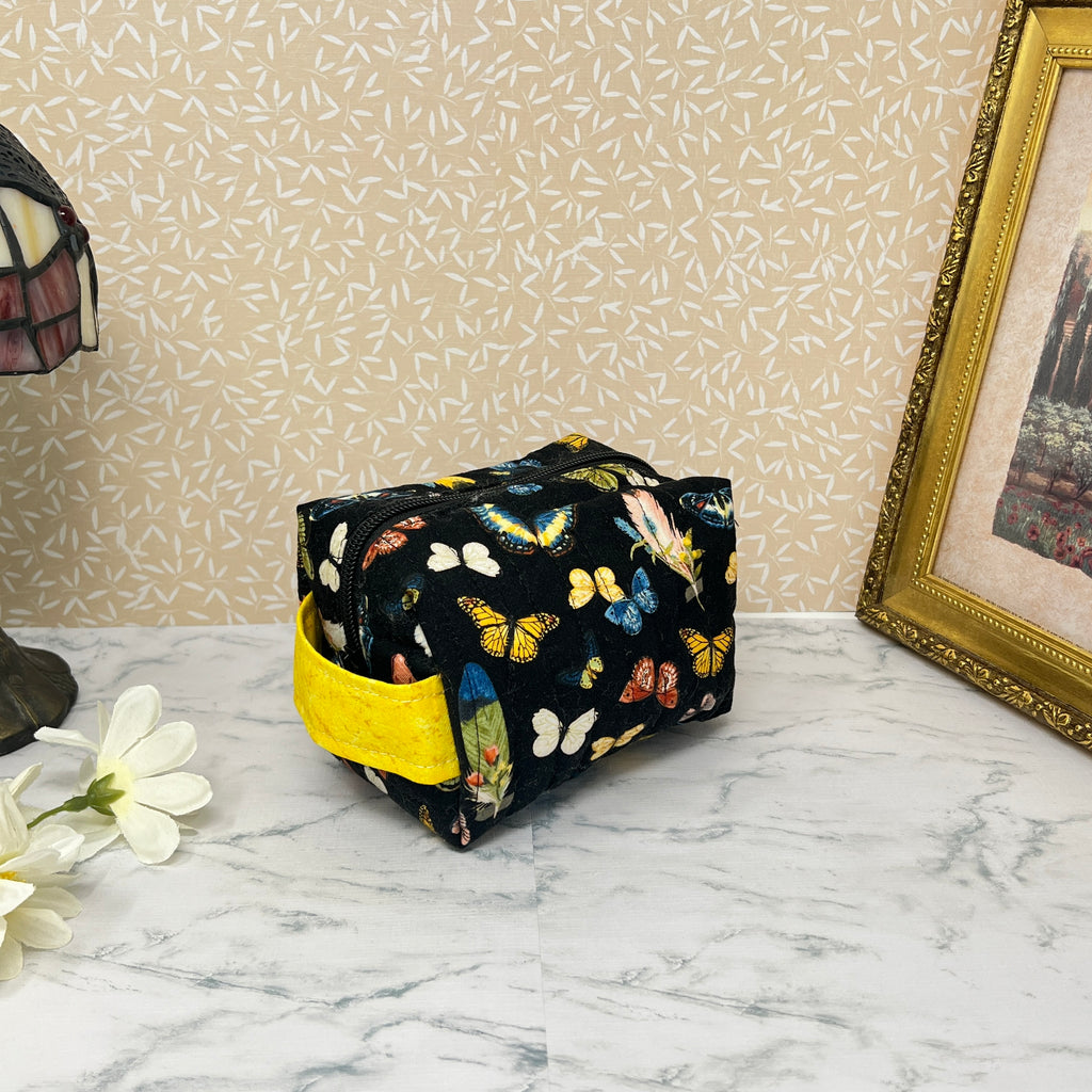 small quilted cosmetic bag with zipper and handle. Fabric is a colorful assortment of butterflies and feathers on a black background with a yellow handle and liner