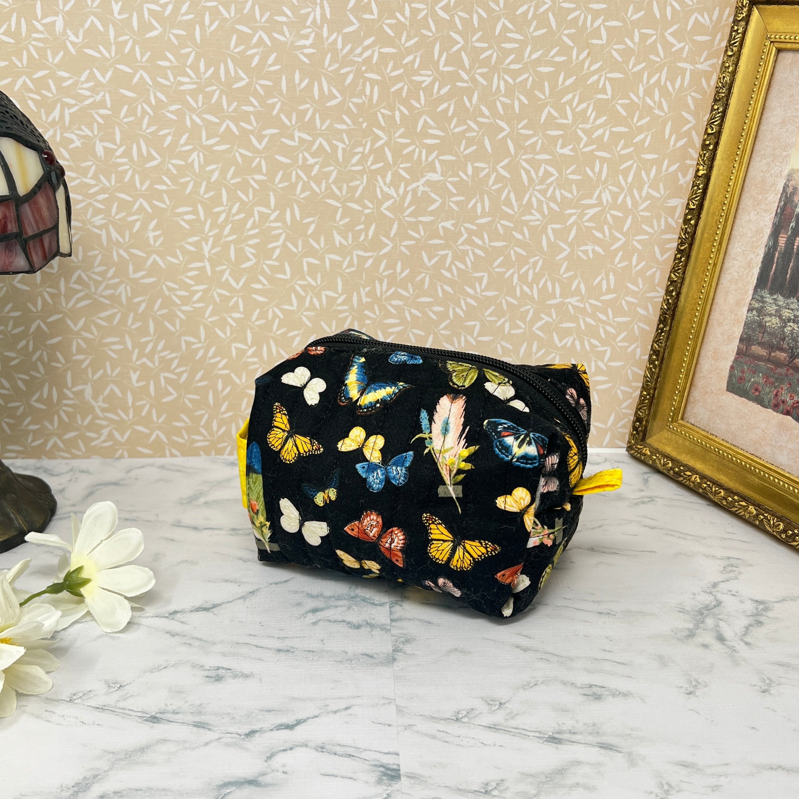 small quilted cosmetic bag with zipper and handle . Fabric is a colorful assortment of butterflies and feathers on a black background with a yellow handle and liner