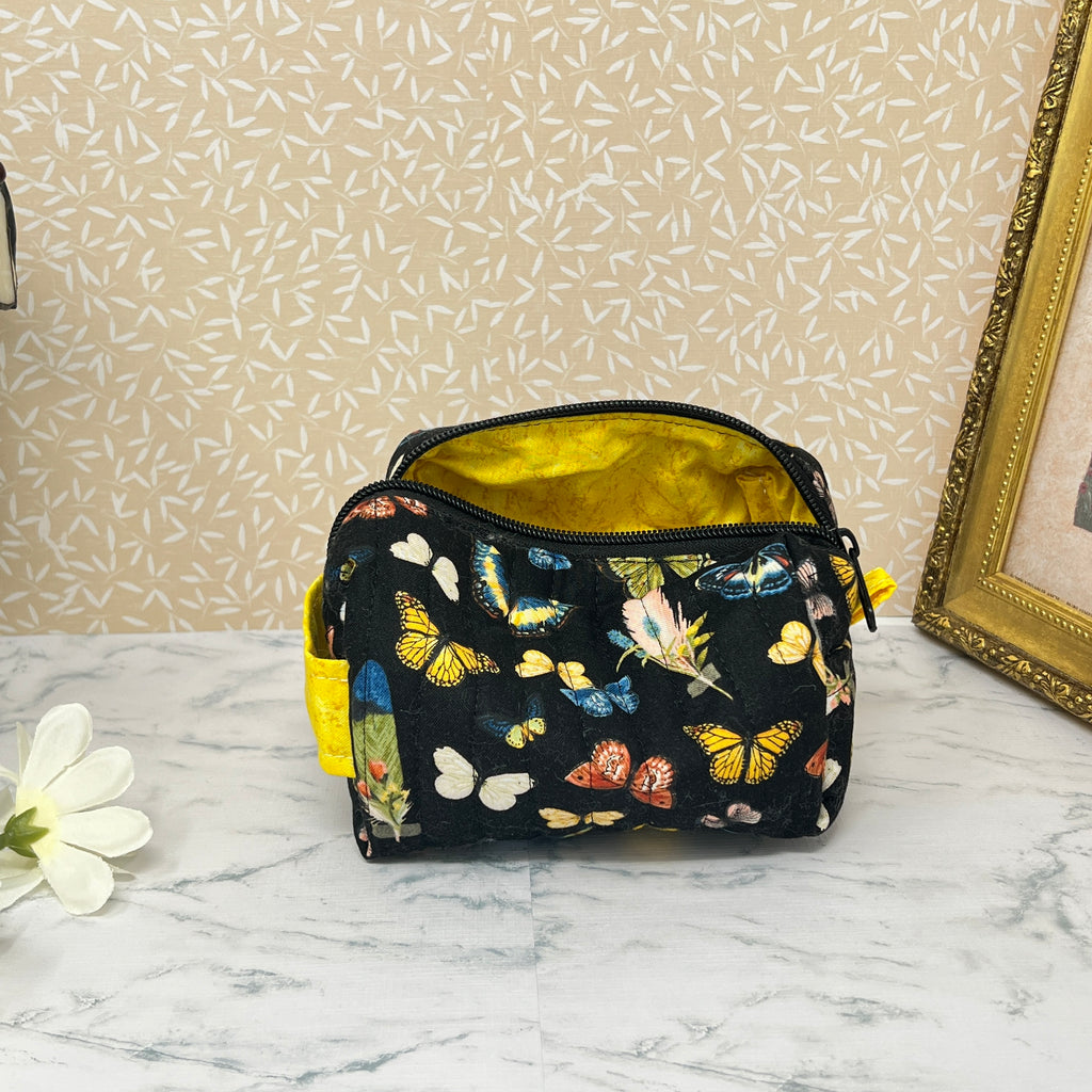 small quilted cosmetic bag with zipper and handle showing the yellow interior lining. Fabric is a colorful assortment of butterflies and feathers on a black background with a yellow handle and liner