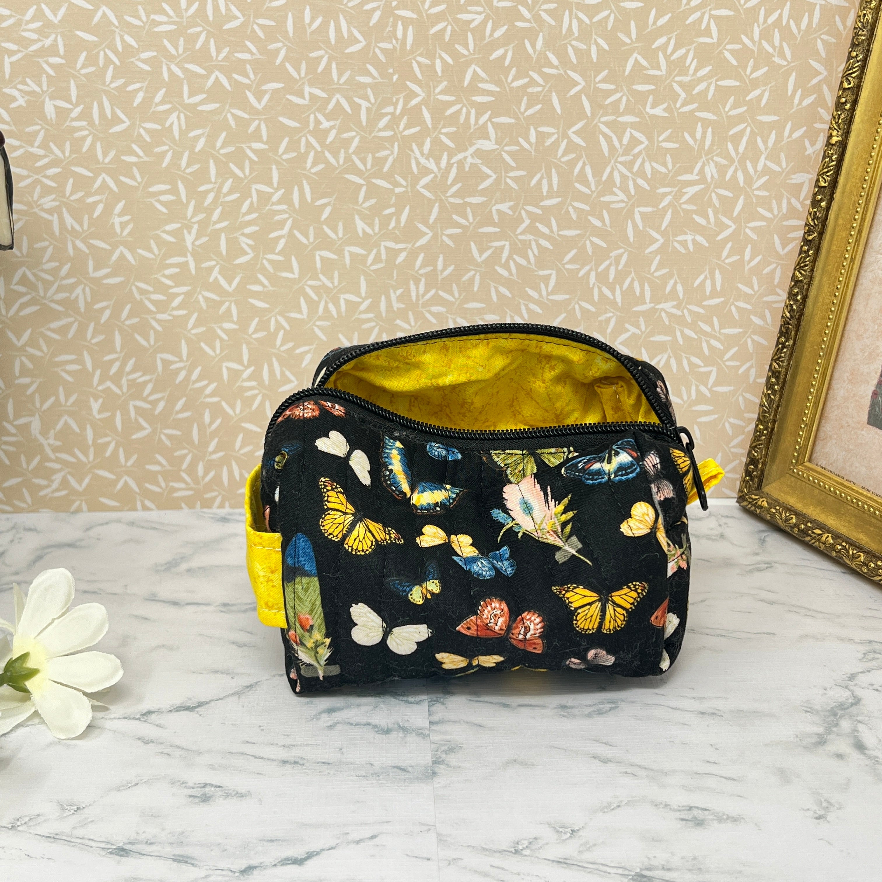 small quilted cosmetic bag with zipper and handle showing the yellow interior lining. Fabric is a colorful assortment of butterflies and feathers on a black background with a yellow handle and liner