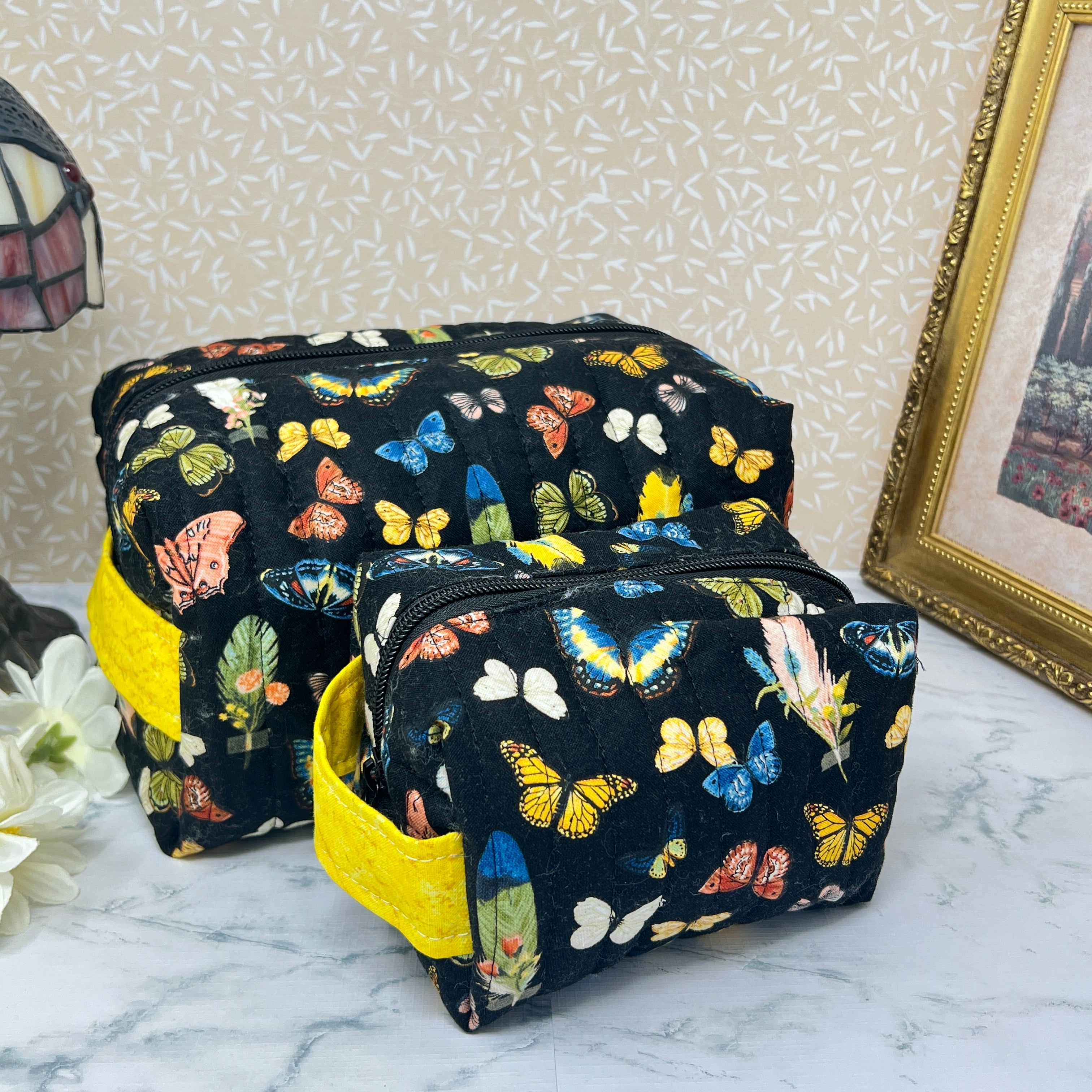 A large and a small quilted cosmetic bag set with zipper and handle.  Also known as a makeup bag or toiletry bag or pouch. Fabric is a colorful assortment of butterflies and feathers on a black background with a yellow handle.