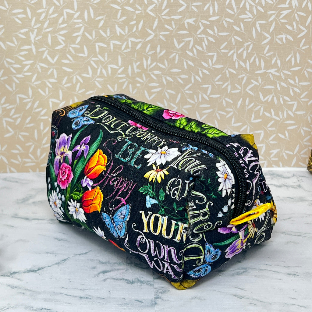 small quilted boxy bag with zipper and handle. Fabric is colorful flowers and positive affirmation text on a black background. Yellow handle and yellow liner. Text; grown your own way, don't worry be happy,