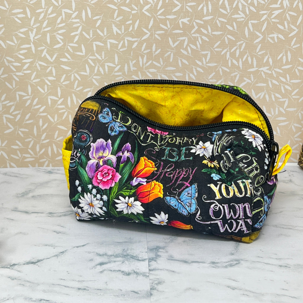small quilted boxy bag with zipper and handle. Fabric is colorful flowers and positive affirmation text on a black background. Yellow handle and yellow liner. Text; grown your own way, don't worry be happy, 