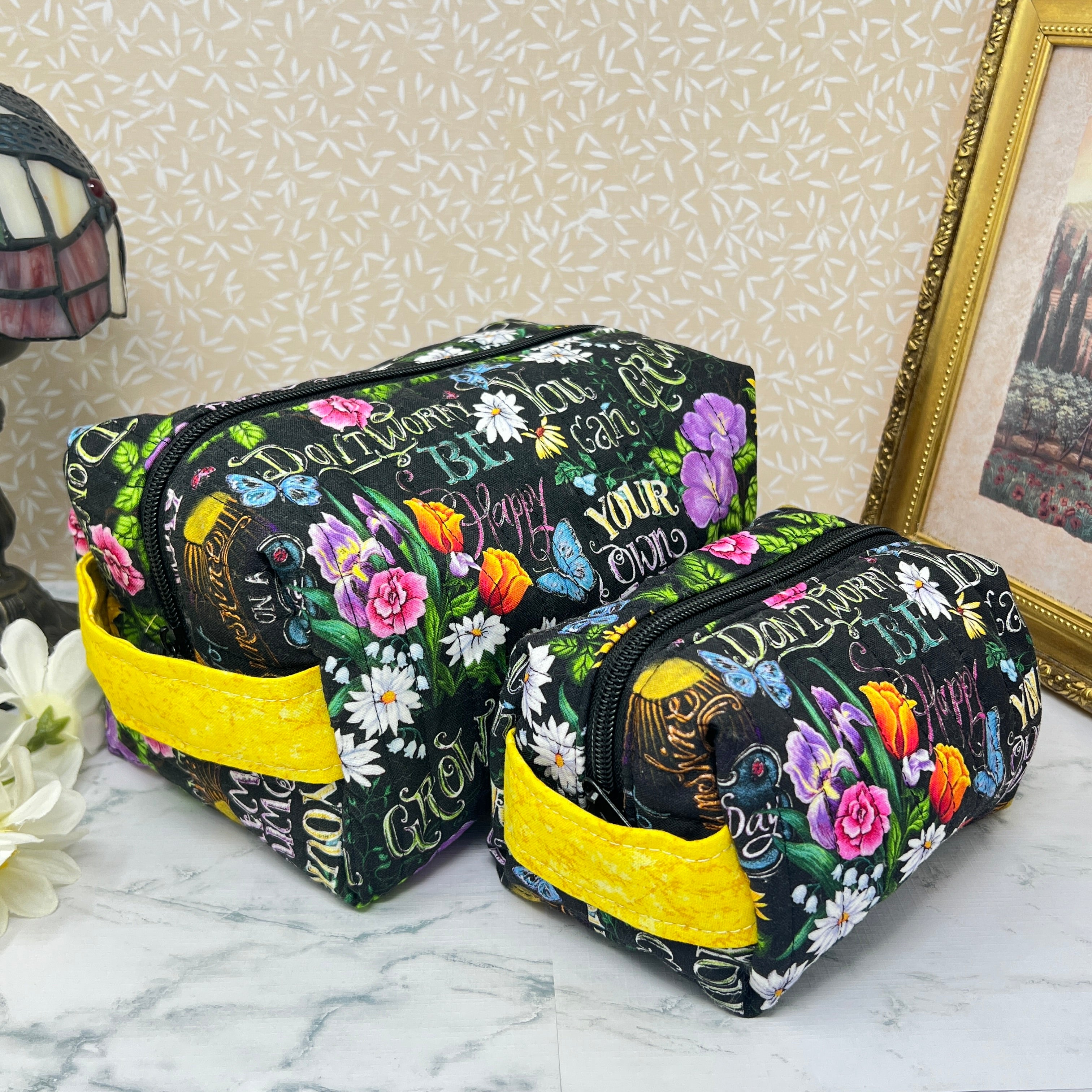 A Large and small quilted boxy bags with zipper and handle. Fabric is colorful flowers and positive affirmation text on a black background. Yellow handle and yellow liner. Text; grown your own way, don't worry be happy,