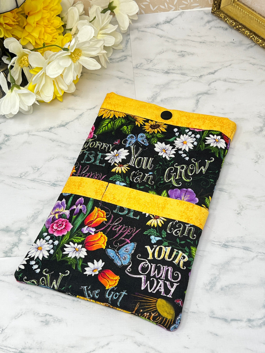 A fabric e-reader sleeve or organizer pouch with a black background, colorful floral print, and motivational text like 'You can GROW YOUR own WAY and 'BE Happy" "Don't Worry". It has bright yellow trim and two front pckets for accessories.
