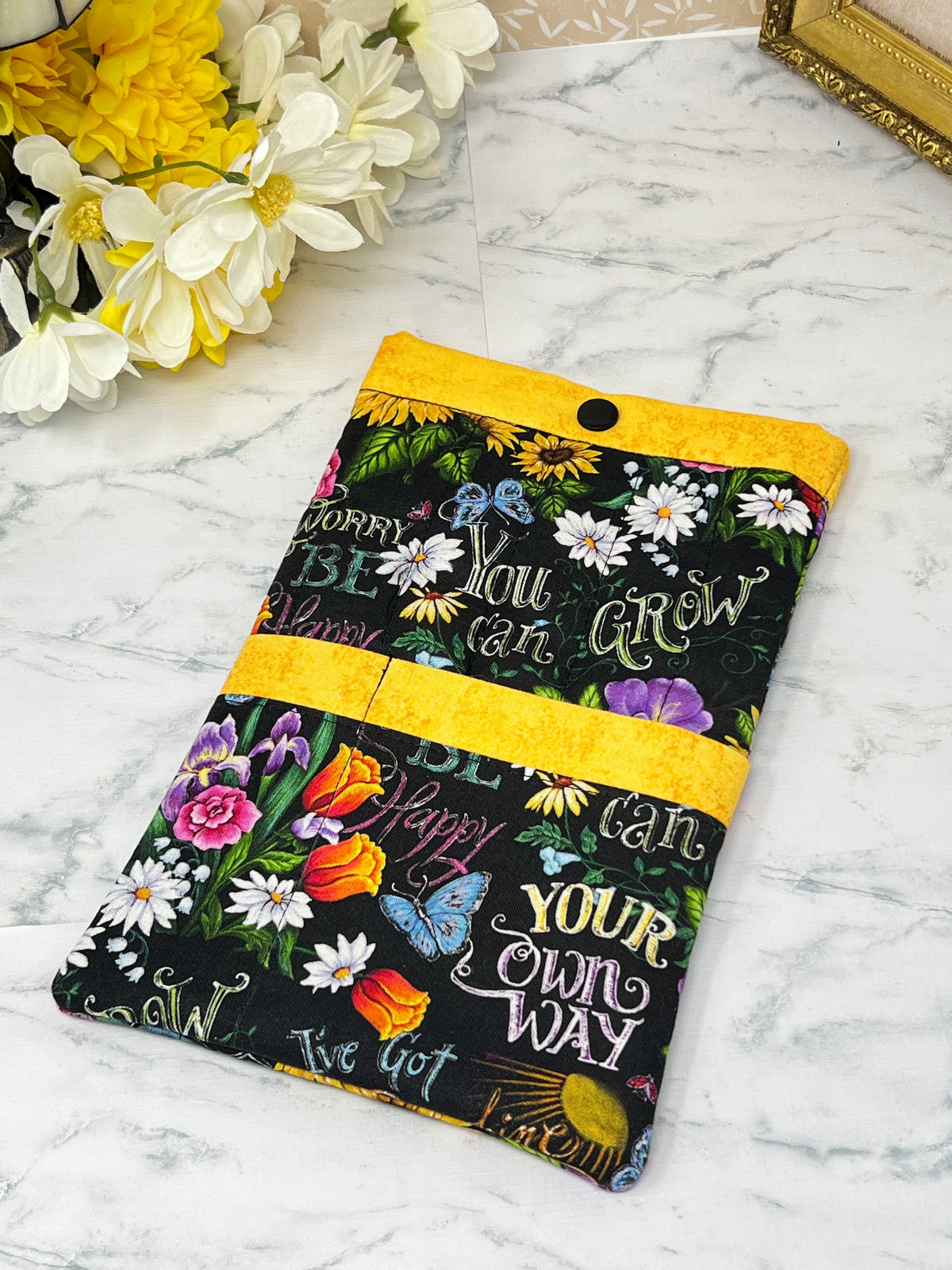 A fabric e-reader sleeve or organizer pouch with a black background, colorful floral print, and motivational text like 'You can GROW YOUR own WAY and 'BE Happy" "Don't Worry". It has bright yellow trim and two front pckets for accessories.
