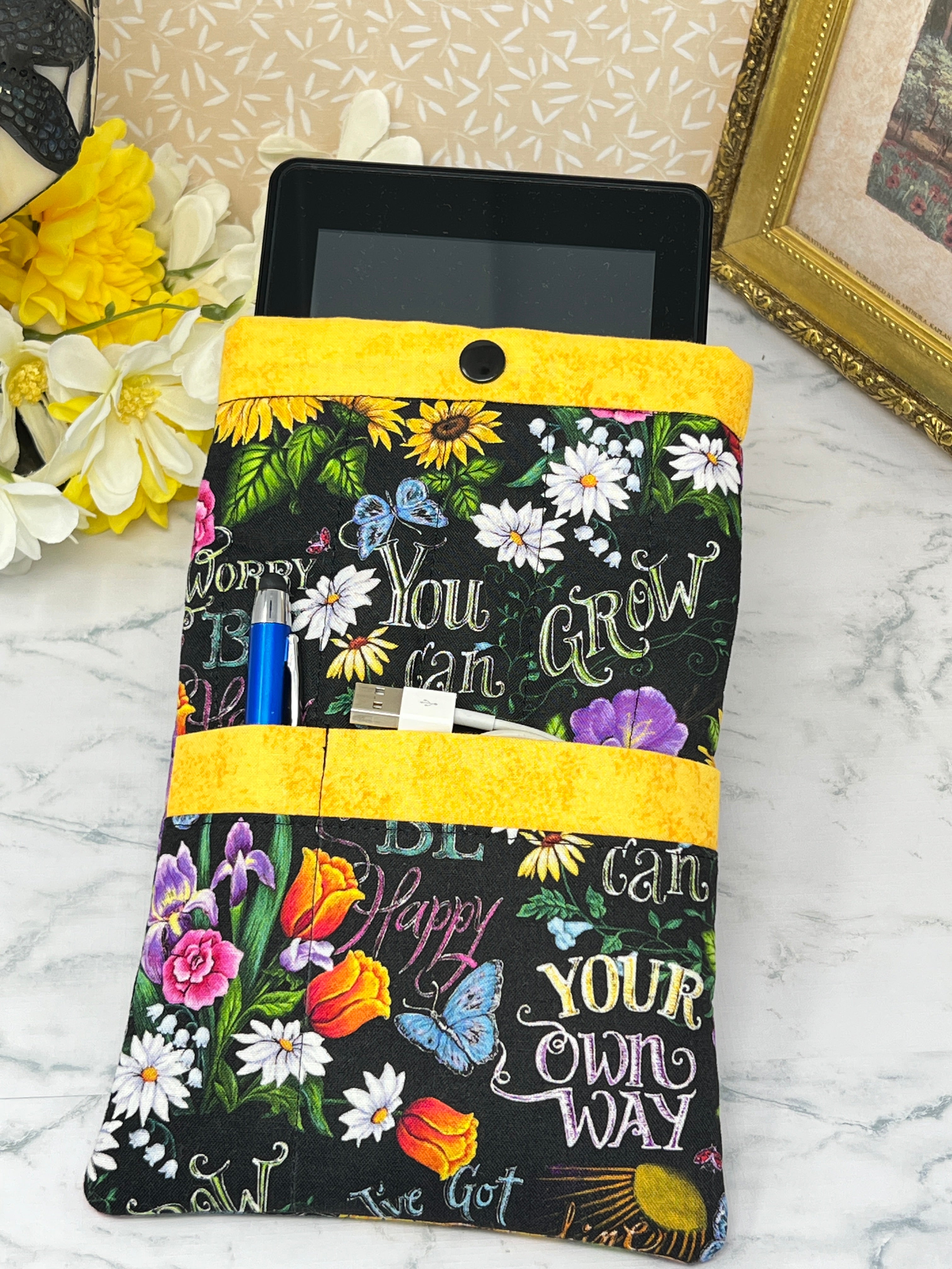 A fabric e-reader sleeve or organizer pouch with a black background, colorful floral print, and motivational text like 'You can GROW YOUR own WAY and 'BE Happy" "Don't Worry". It has bright yellow trim and is shown holding a blue pen stylus and charging cord in the front pockets and an E-reader in the main pocket