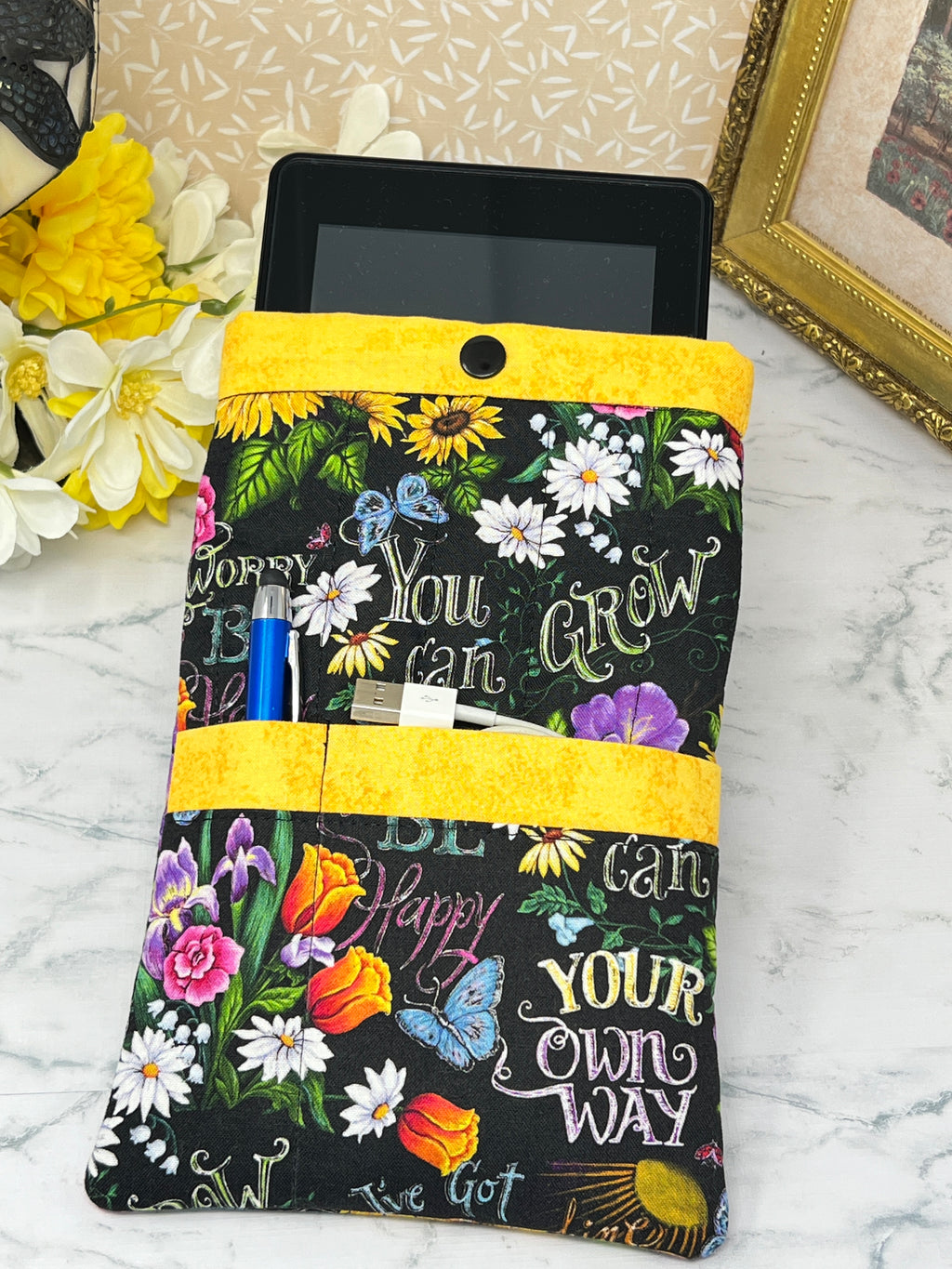 A fabric e-reader sleeve or organizer pouch with a black background, colorful floral print, and motivational text like 'You can GROW YOUR own WAY and 'BE Happy" "Don't Worry". It has bright yellow trim and is shown holding a blue pen stylus, charging cords in the front pockets and an E-reader in the main pocket