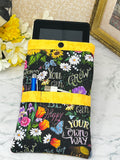 A fabric e-reader sleeve or organizer pouch with a black background, colorful floral print, and motivational text like 'You can GROW YOUR own WAY and 'BE Happy" "Don't Worry". It has bright yellow trim and is shown holding a blue pen stylus, charging cords in the front pockets and an E-reader in the main pocket
