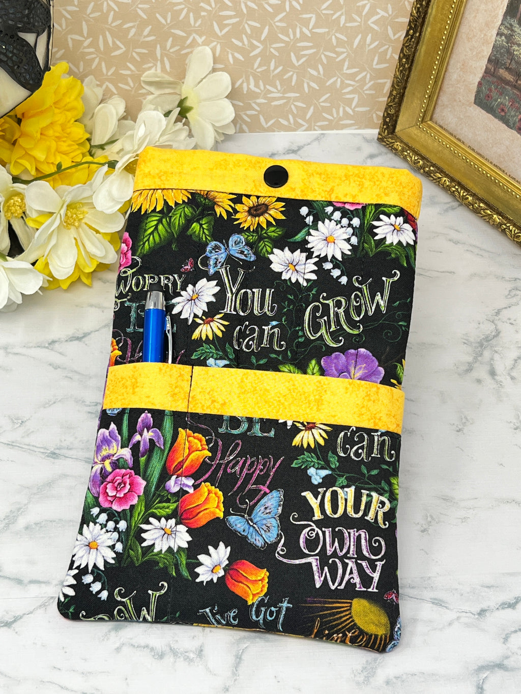 A fabric e-reader sleeve or organizer pouch with a black background, colorful floral print, and motivational text like 'You can GROW YOUR own WAY and 'BE Happy" "Don't Worry". It has bright yellow trim and is shown holding a blue pen stylus, in one of the front pockets.