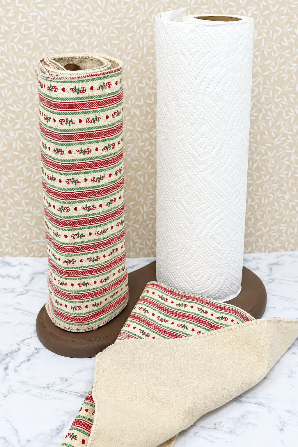 A roll of textured white paper towels standing next to a roll of cream-colored ribbon with red and green holiday stripes, both displayed on a dark brown dual towel stand on a marble surface.